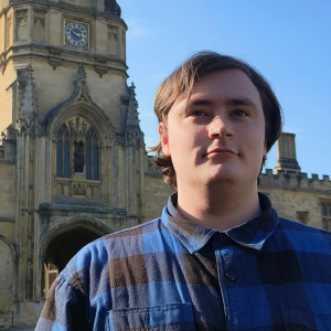 Young man with a blue shirt stood in front of a large old-fashioned building with a big clock.