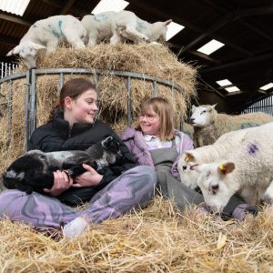 A young adult and a child petting a lamb whilst sitting down on hay.