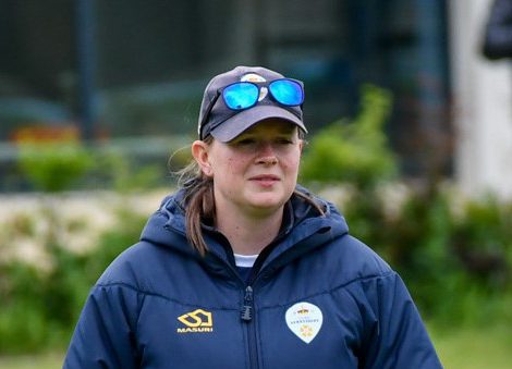 Female cricket coach, Emma Thatcher, wearing a blue puffer jacket and a baseball cap.