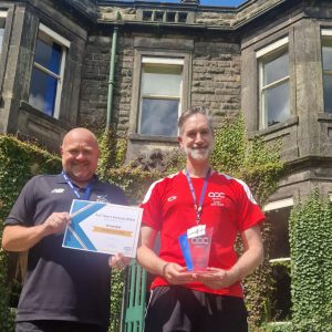Two men wearing sports gear whilst holding an AoC Award and Certificate in front of a large brick building.