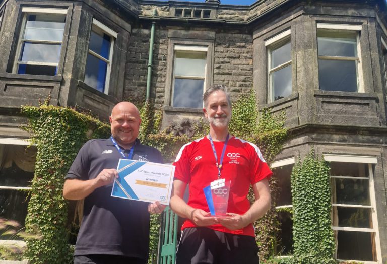 Two men wearing sports gear whilst holding an AoC Award and Certificate in front of a large brick building.