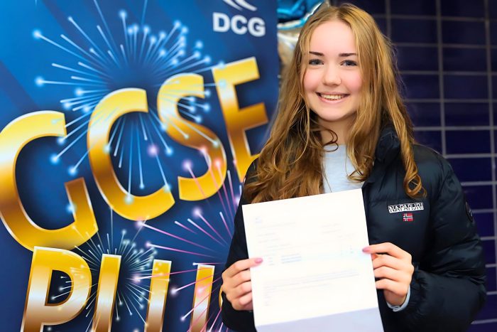 Young lady with brown hair smiling whilst holding a piece of paper.