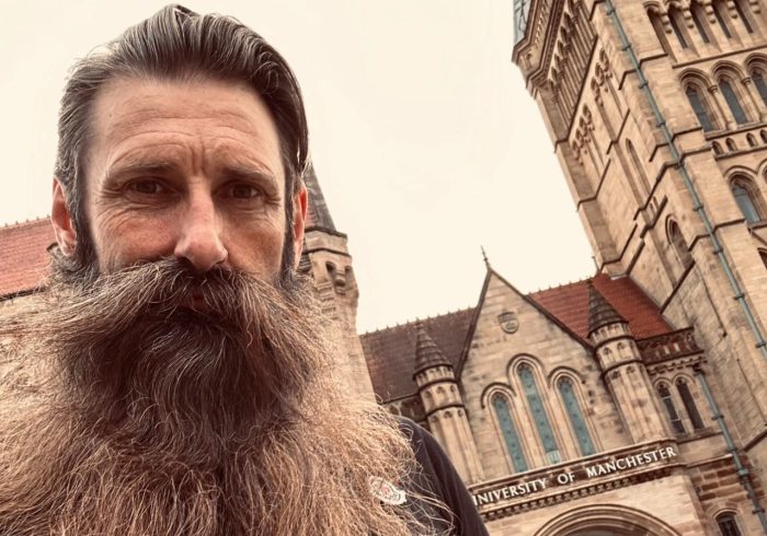 Man with a beard stood in front of an old-fashioned brick building.