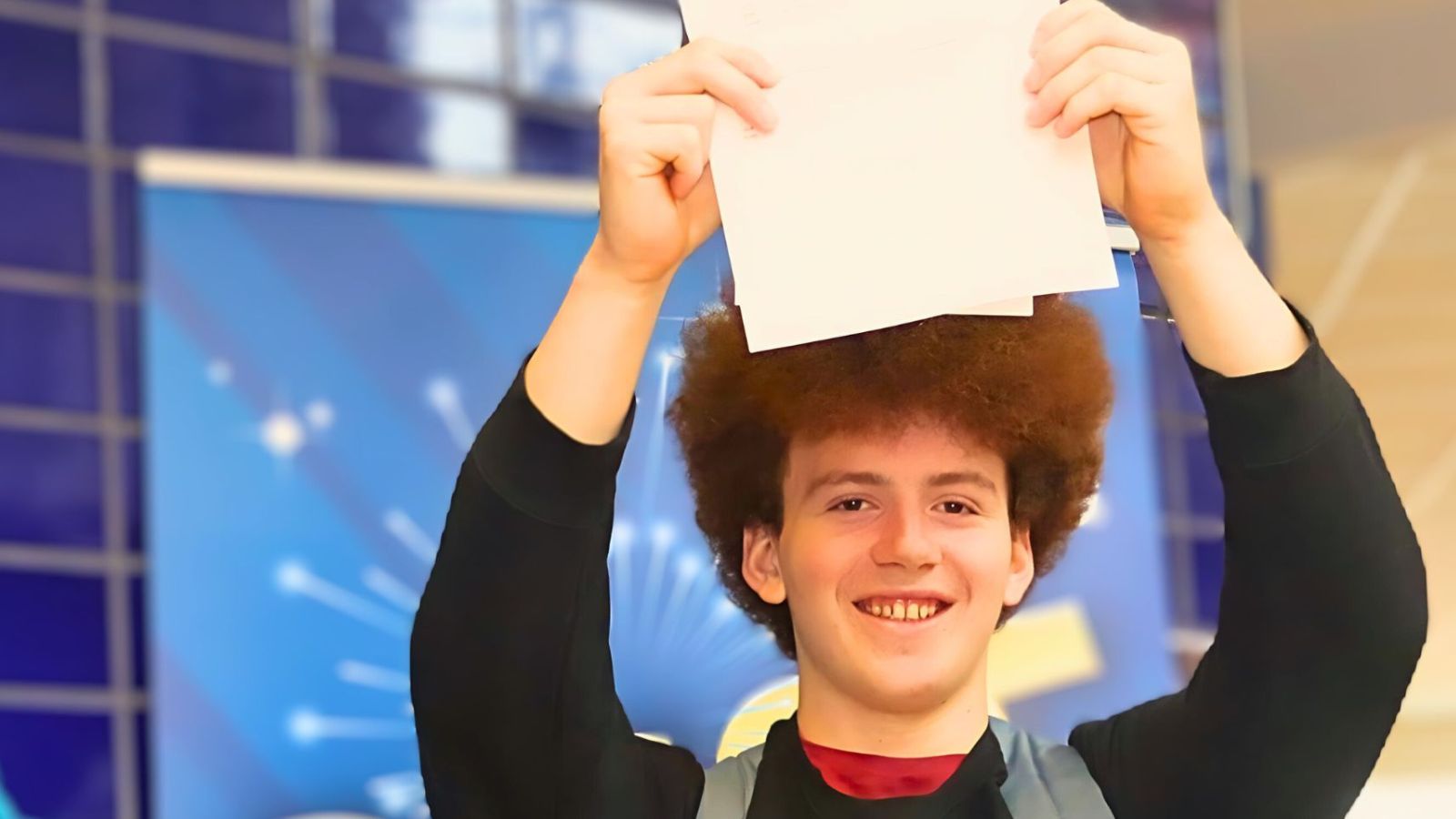 Young man with curly hair holding a peice of paper aloft.