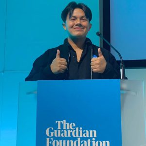 A young man stood on a blue podium that reads ' The Guardian Foundation' delivering a presentation