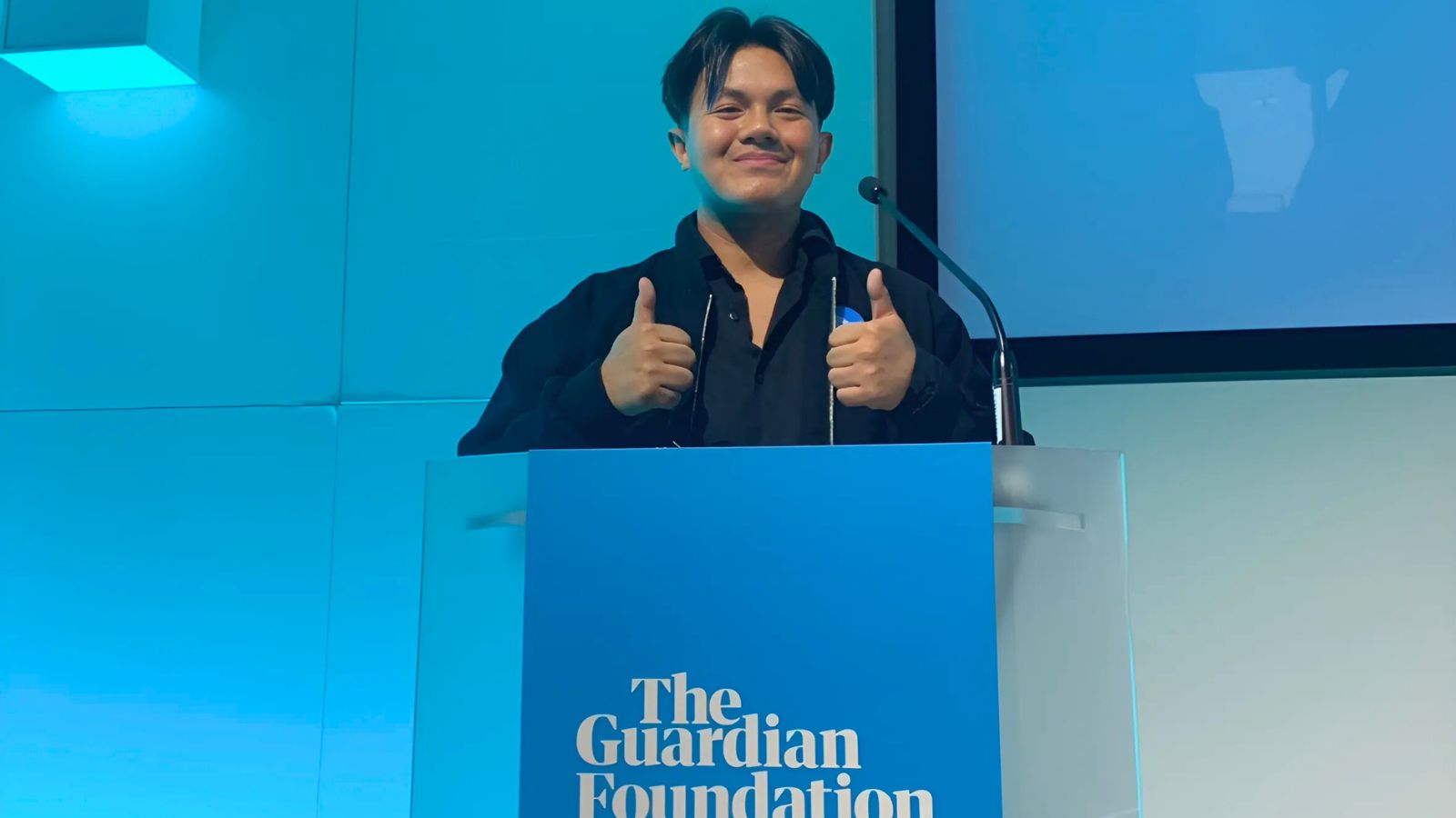 A young man stood on a blue podium that reads ' The Guardian Foundation' delivering a presentation