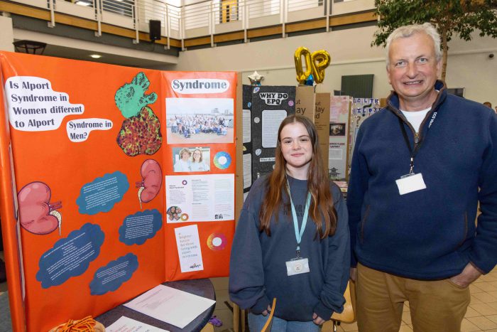 A young Derby College student stood by her presentation, an orange background with various pictures and text.