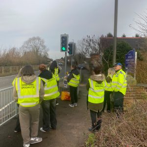 A group of young students wearing hi-viz jackets stood by a traffic light.