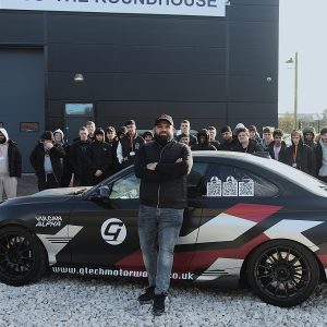 Man stood in front of a car with arms folded, a group of students stood behind the car.