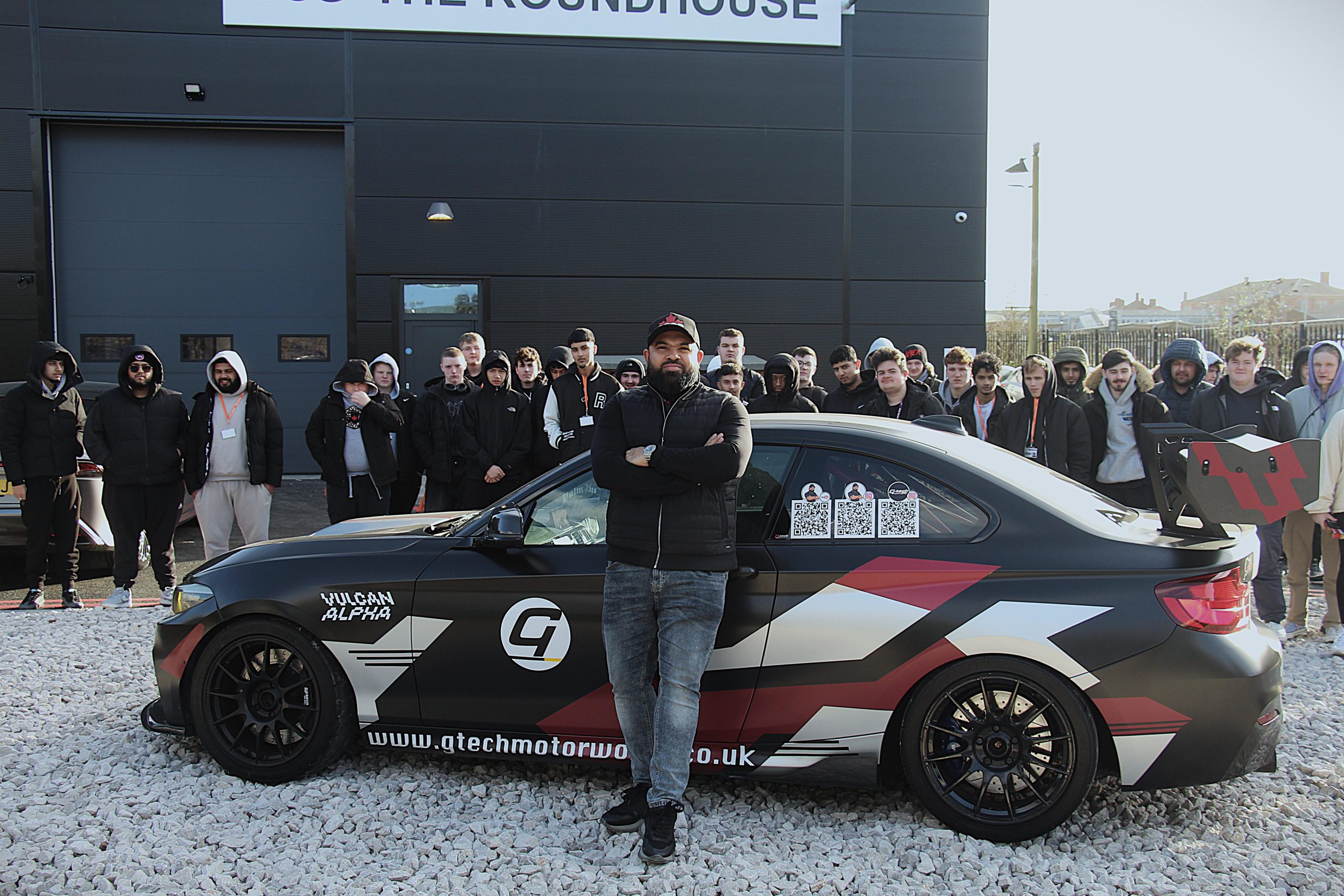 Man stood in front of a car with arms folded, a group of students stood behind the car.