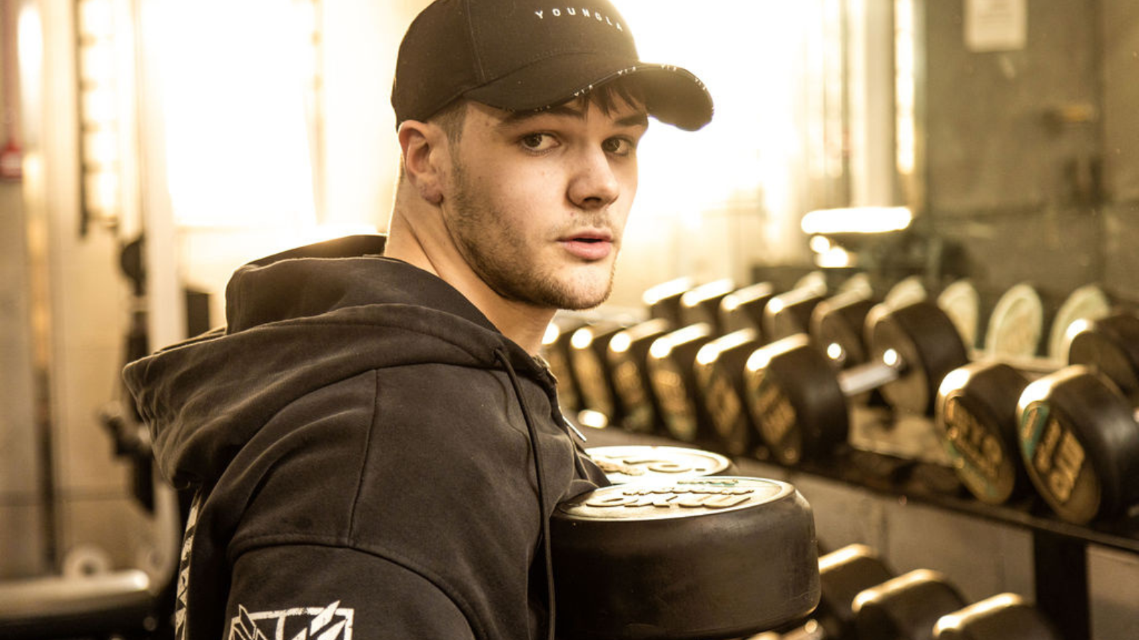 Young man wearing a black hoodie and baseball cap lifting dumbells