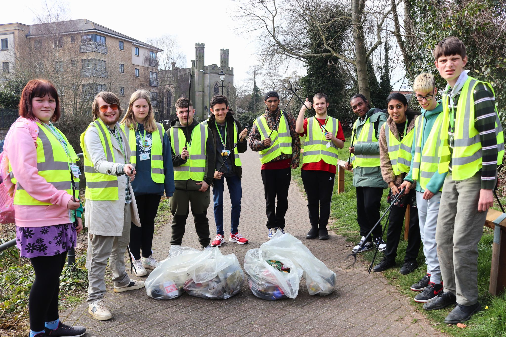 A group of students wearing Hi-Viz vests holding litter pickers, stood in front of full bin bags.