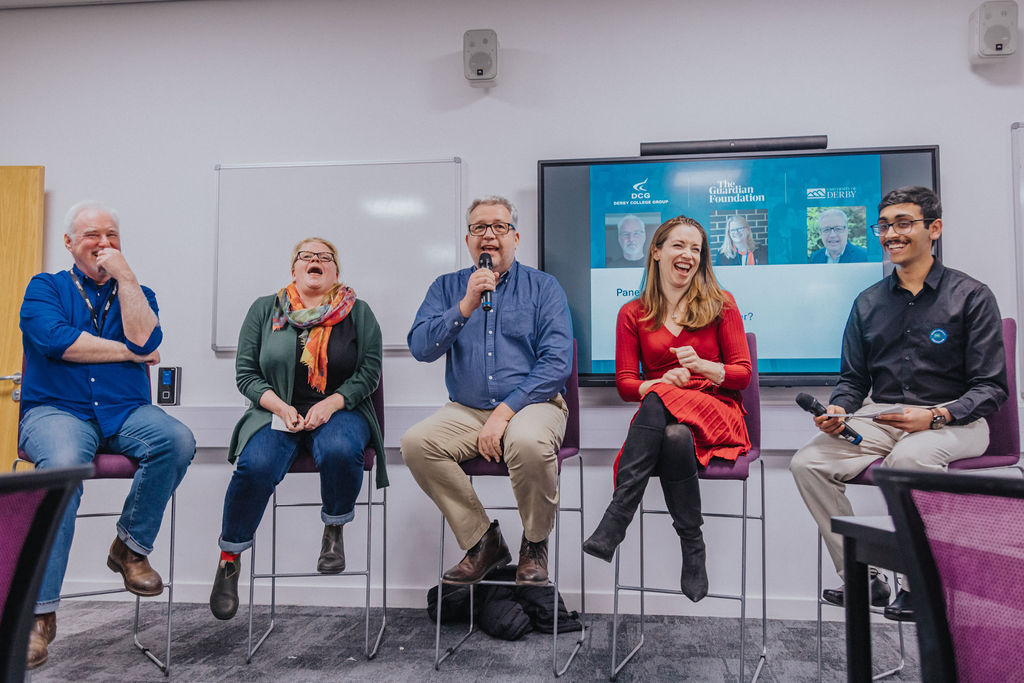 Five speakers sat on stools in front of a screen holding microphones.